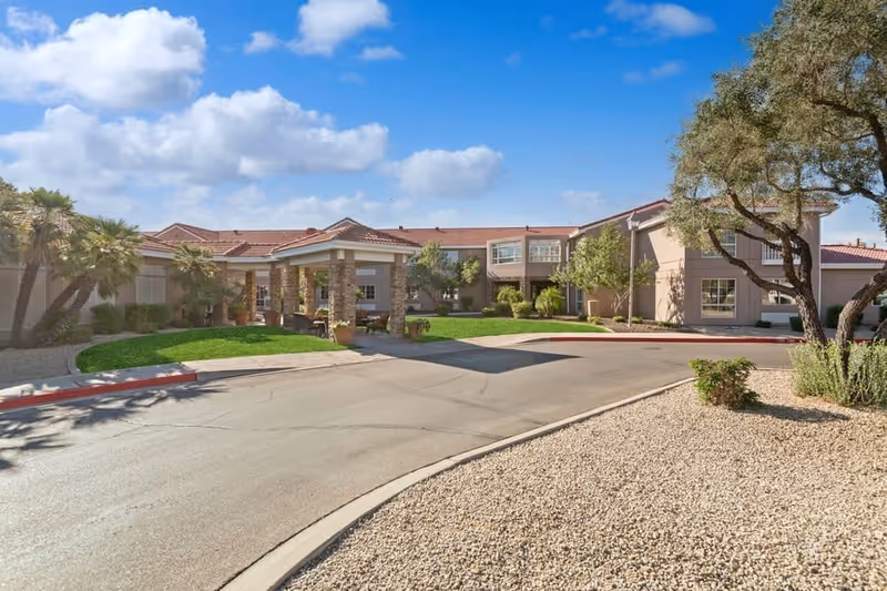 Exterior view of The Gardens Of Scottsdale senior living facility showing a driveway leading to the main entrance with a covered drop-off area, surrounded by landscaped greenery, palm trees, and desert plants under a blue sky with scattered clouds.
