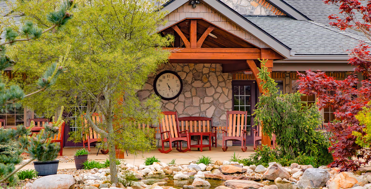 Outdoor patio area at The Inn at Library Way featuring wooden rocking chairs and a bench under a covered porch with a stone wall and a large clock. The area is surrounded by lush greenery, trees, and a rock-lined pond in the foreground.