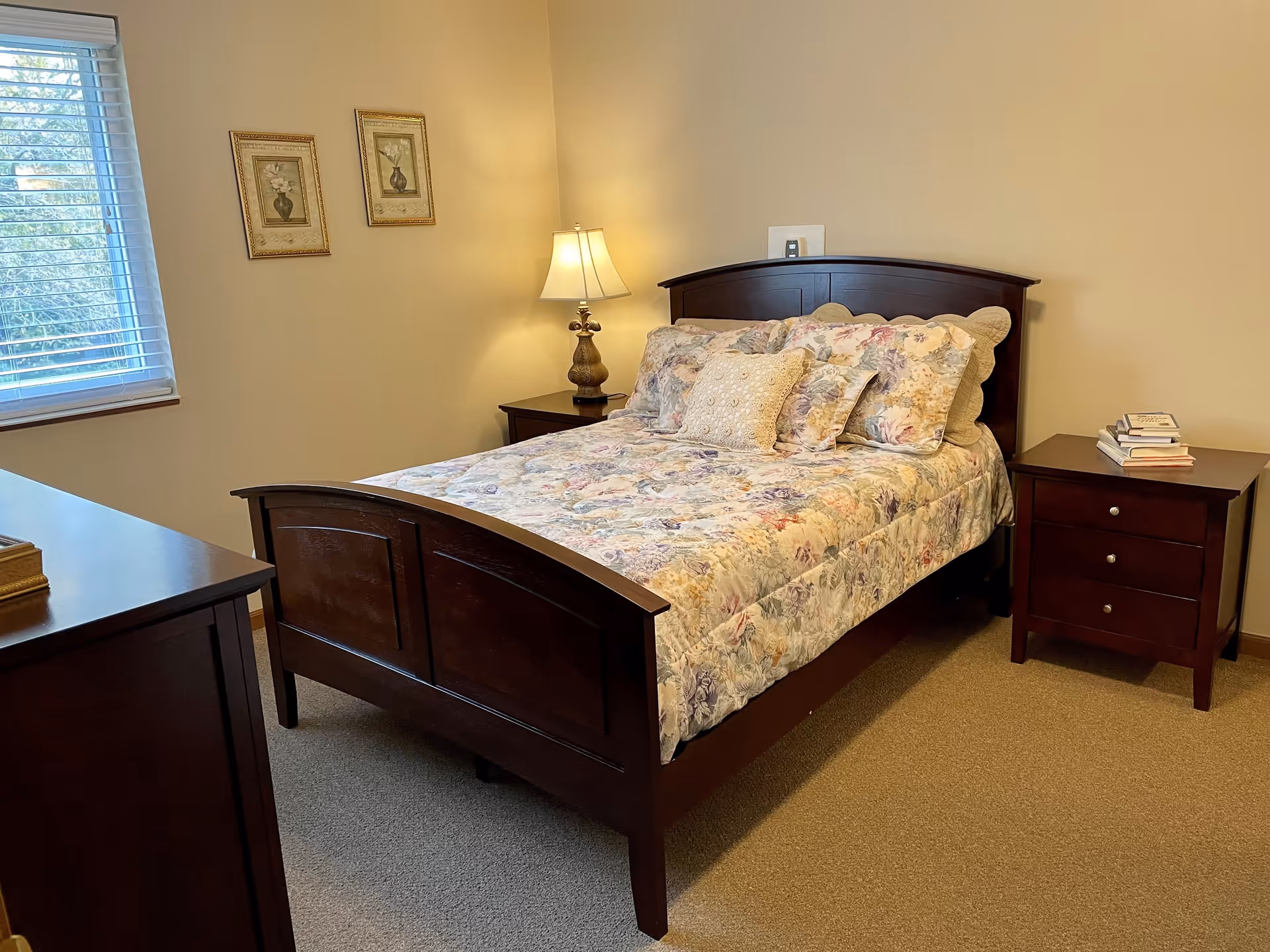 A bedroom with a wooden bed frame and floral bedding, two matching wooden nightstands with a lamp on one and a stack of books on the other, beige walls with two framed pictures, and a window with blinds letting in natural light.