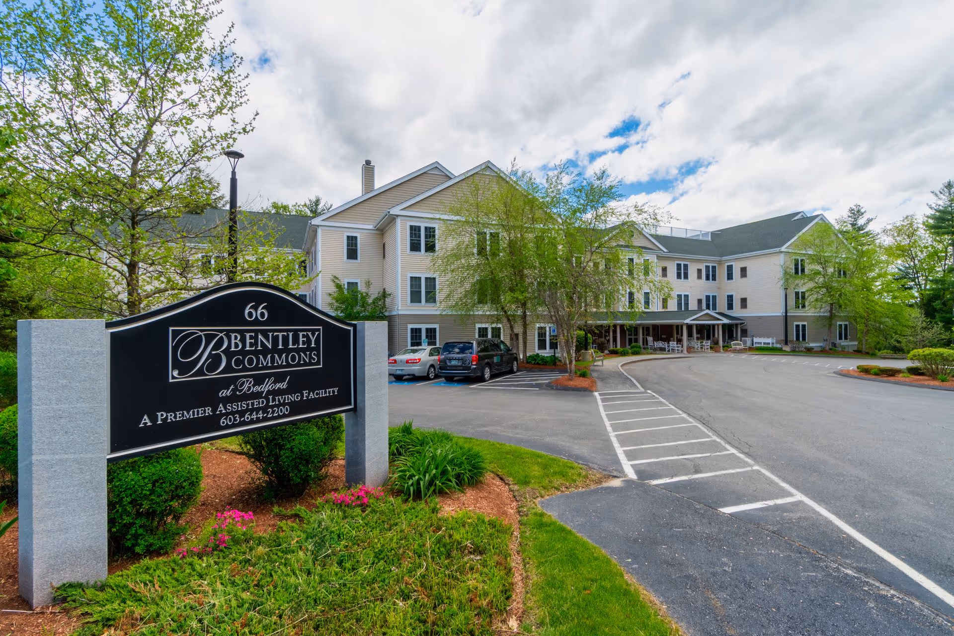Exterior view of Bentley Commons at Bedford, a premier assisted living facility, showing a large beige multi-story building with several windows, a driveway, parked cars, and a landscaped area with green grass, bushes, and flowers under a partly cloudy sky.