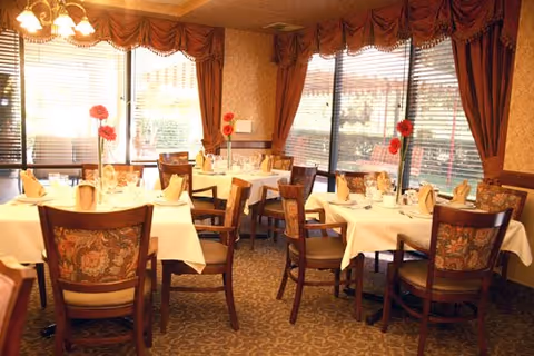 A warmly lit dining room with several tables covered in white tablecloths, each set with folded napkins, glassware, and plates. The chairs have floral upholstery, and large windows with blinds and brown curtains allow natural light to enter. Red flowers in vases decorate the tables.