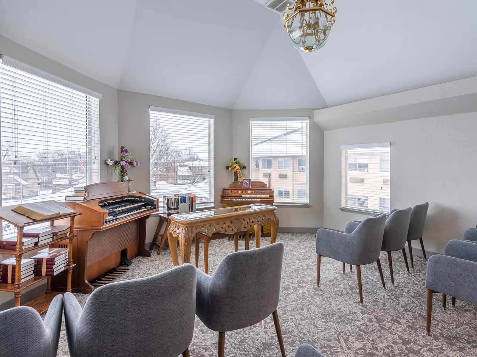 Bright common room with an electric organ and wooden piano by large windows and rows of gray chairs.