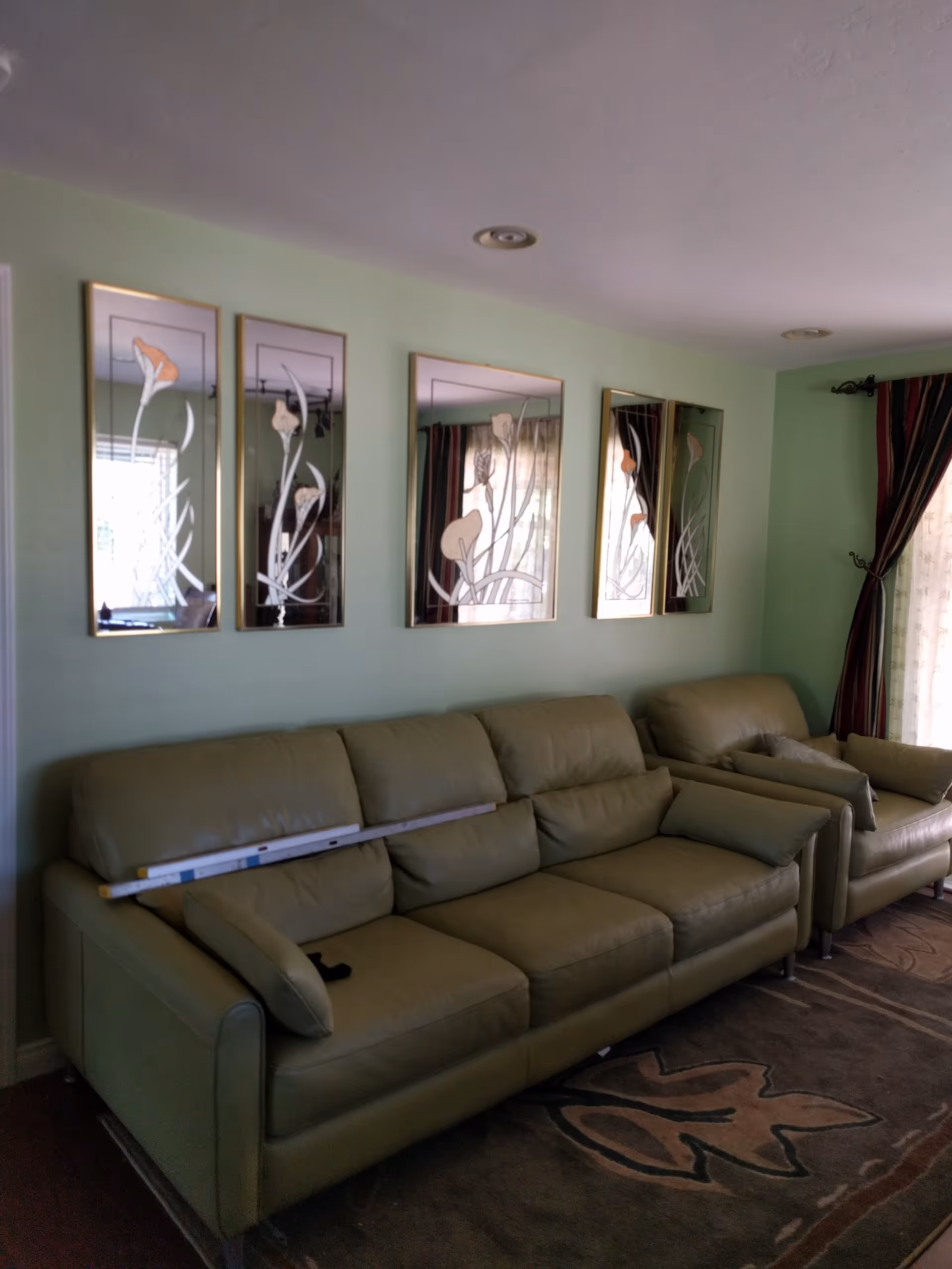 A living room with a green leather sofa and matching armchair against a light green wall. Above the sofa are five decorative mirrors with floral designs. A patterned rug is on the floor, and a window with dark curtains is visible on the right side.