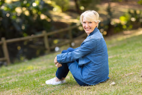 A smiling elderly woman with short blonde hair wearing a blue denim jacket and dark pants sits on grass outdoors with a wooden fence and trees in the background.