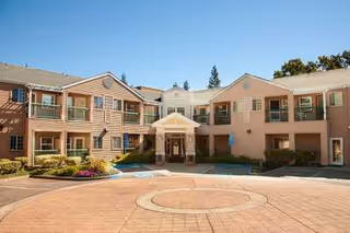 Exterior view of a two-story senior living facility building with beige siding and multiple windows. The entrance has a covered porch with white columns, and there is a circular driveway in front with landscaped bushes and flowers. Trees and a clear blue sky are visible in the background.