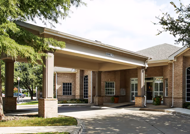 Entrance area of a senior living facility with a covered drop-off zone supported by columns, brick exterior walls, large windows, and surrounding greenery including trees and grass.
