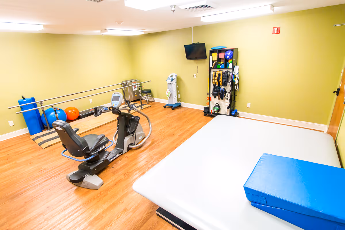 A bright therapy/exercise room with a recumbent bike, parallel bars, exercise balls, mats, and a wall-mounted TV.