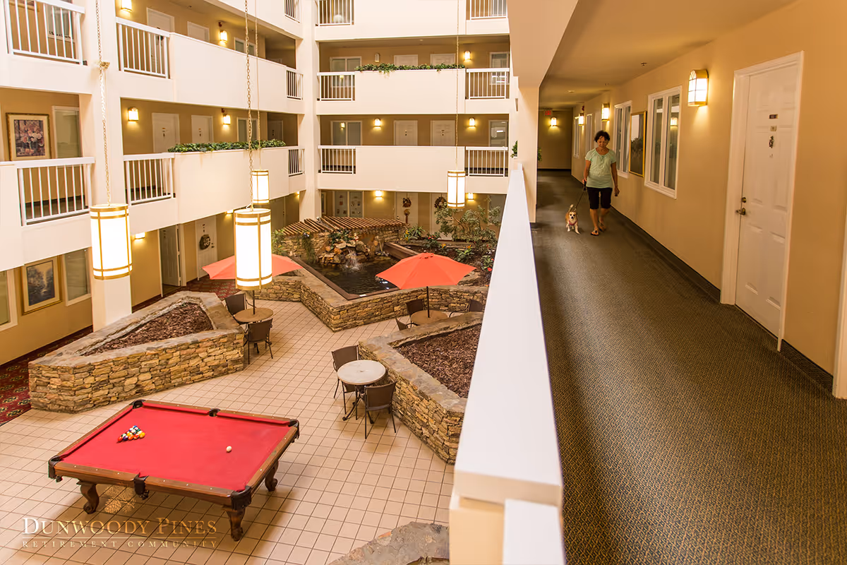 Interior atrium of a retirement community with a red pool table, seating areas, indoor planters and a woman walking a dog along the hallway.