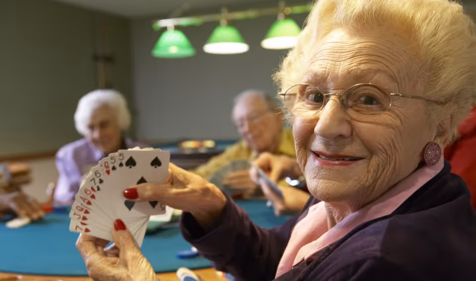 An elderly woman with glasses and a smile is holding a fan of playing cards while sitting at a card table. In the background, other elderly individuals are also engaged in playing cards under green hanging lights.