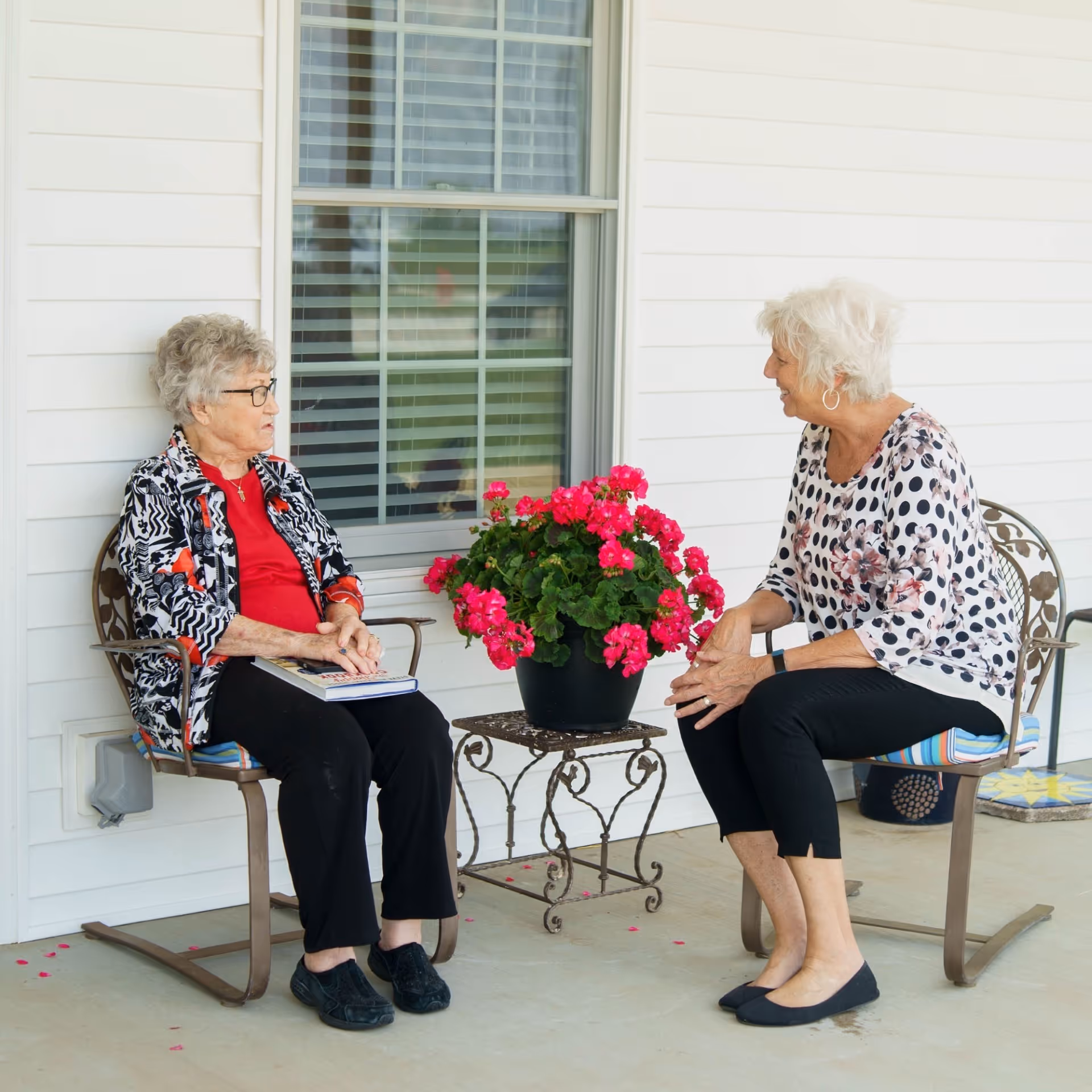Two elderly women sitting on metal chairs on a porch outside a white building, engaged in conversation. There is a small metal table between them with a pot of bright pink flowers. One woman is holding a book on her lap.