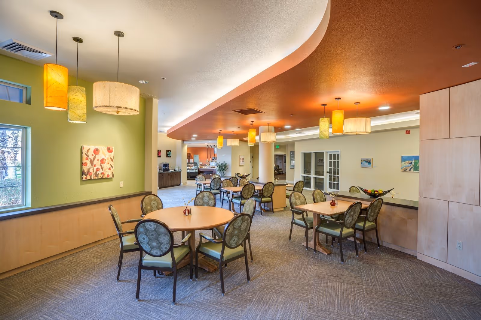 A spacious dining area in a senior living facility with round and square tables surrounded by cushioned chairs. The room features a green accent wall with a floral painting, multiple hanging pendant lights with warm tones, and a curved ceiling design with recessed lighting. In the background, there is a counter area and more seating, with large windows allowing natural light to enter.