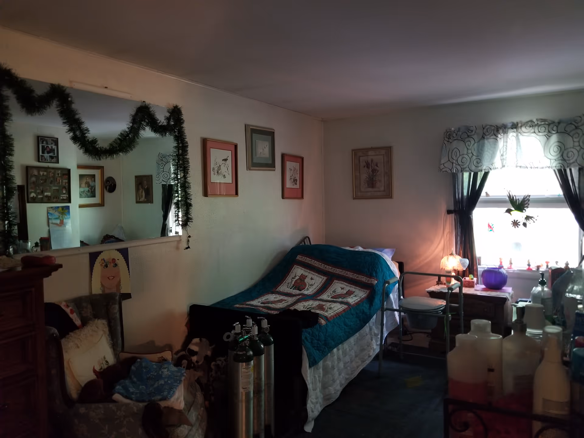 A cozy bedroom with a single bed covered by a blue quilt with a decorative pattern. The room has a large mirror on one wall decorated with green garland, several framed pictures, and a window with sheer curtains letting in natural light. There is a chair with pillows, oxygen tanks near the bed, a small bedside table with a lamp, and various bottles and containers on a surface in the foreground.