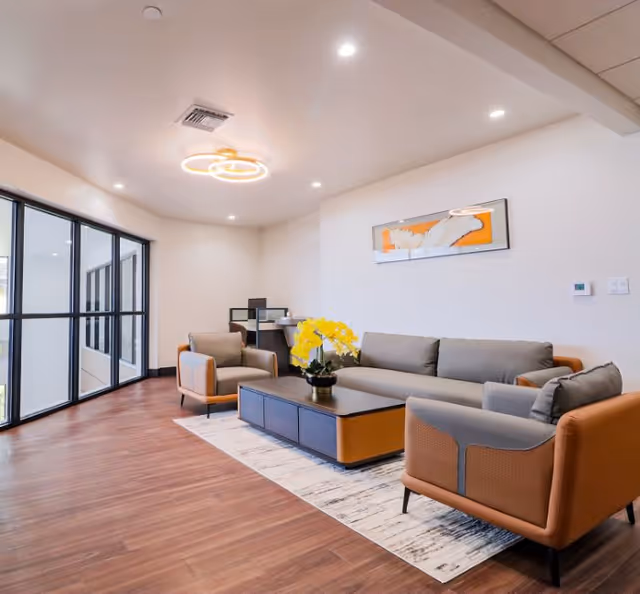 A modern living room with a gray sofa, two matching armchairs with brown accents, a rectangular coffee table with a vase of yellow flowers, a patterned rug, and a large window with black frames letting in natural light. The room has wooden flooring, white walls, recessed ceiling lights, and a contemporary circular ceiling light fixture. A framed abstract artwork is hung on the wall above the sofa.
