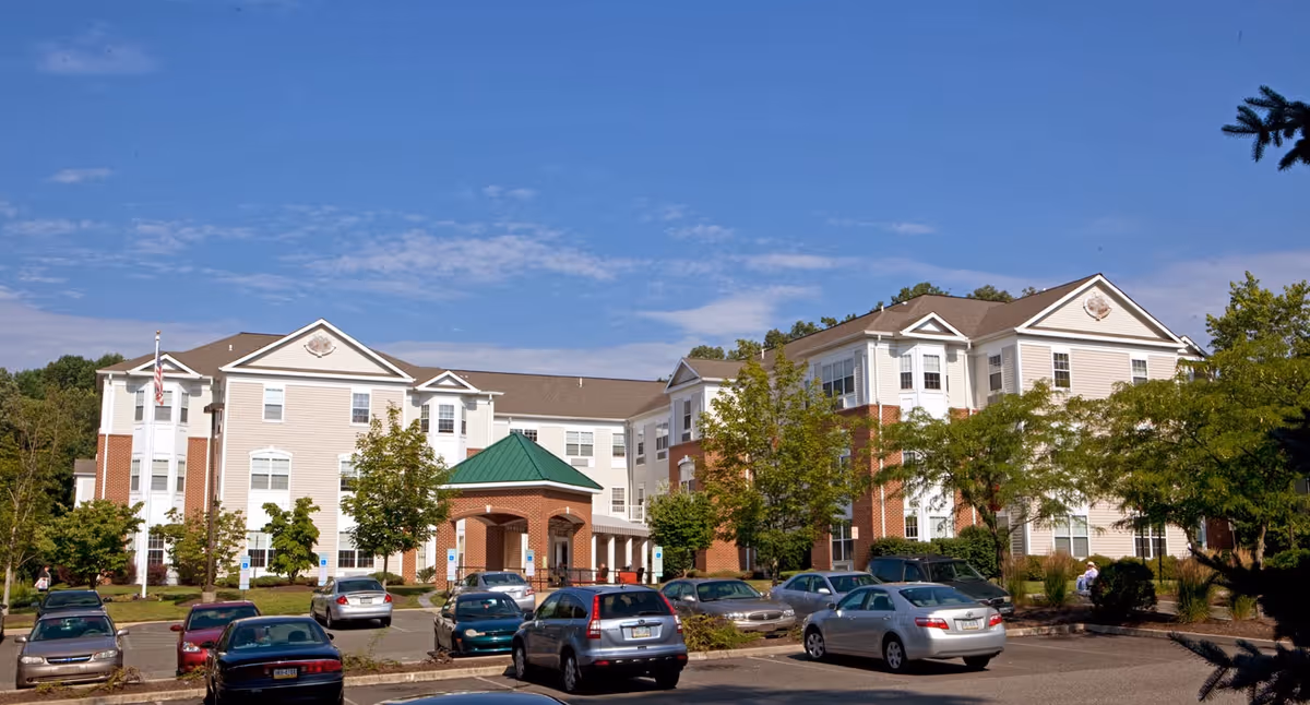 Three-story senior living building with a covered entrance, trees, and a parking lot with cars under a blue sky.