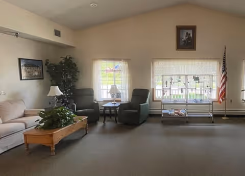 A spacious assisted-living common room with sofas and armchairs around a coffee table, side tables with lamps, windows, and an American flag.