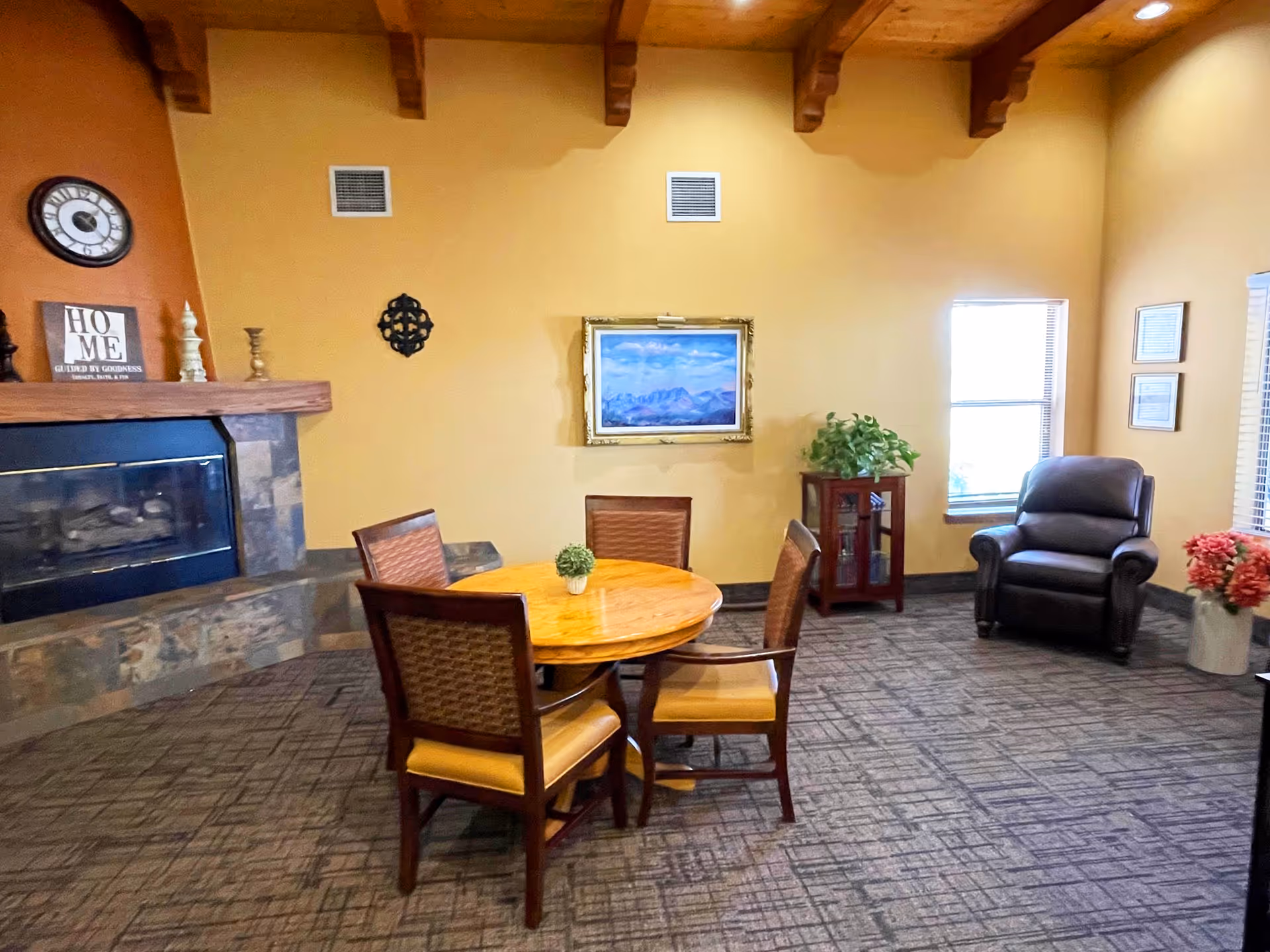 A cozy living room area with a round wooden table surrounded by four chairs, a stone fireplace with a clock and decorative items on the mantel, a framed landscape painting on the wall, a leather armchair near two windows, a small cabinet with a plant on top, and a vase with flowers on the floor.