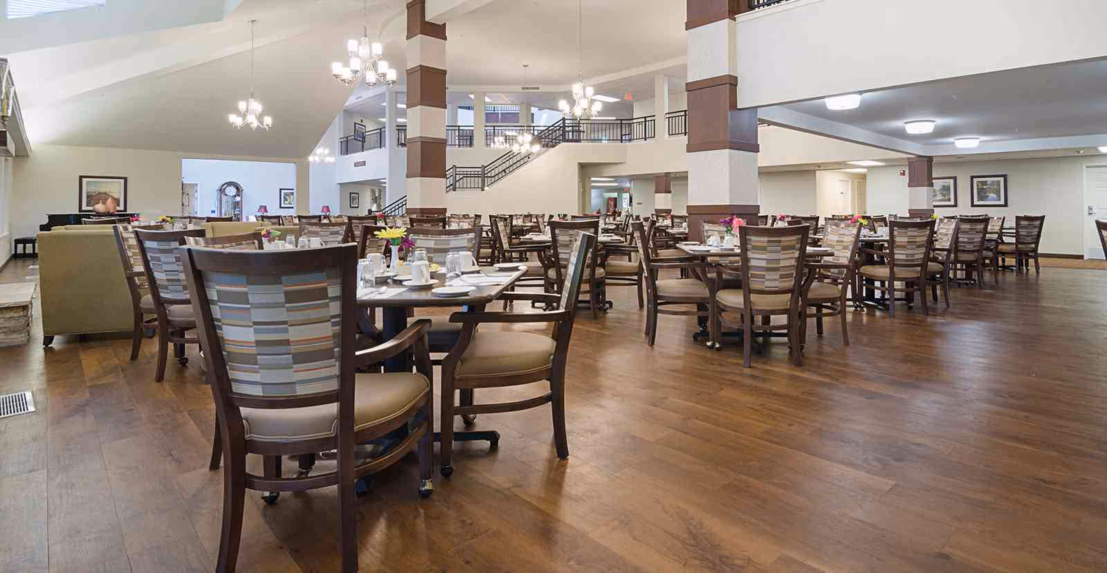 Spacious dining area with multiple wooden tables and chairs arranged neatly. Each table is set with white plates, cups, and small flower vases. The room has wooden flooring, high ceilings with chandeliers, and a staircase leading to an upper level. The walls are decorated with framed pictures.