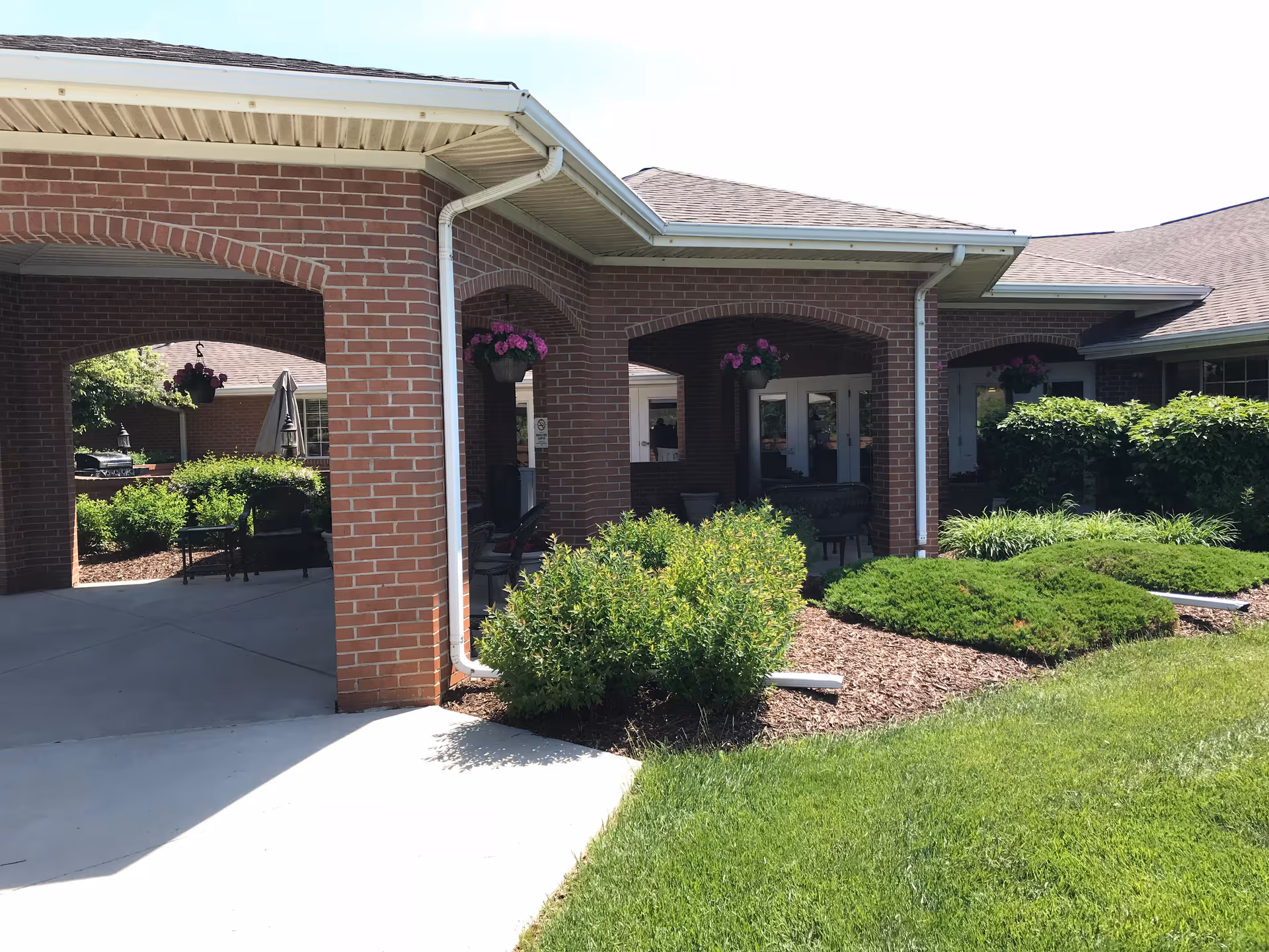 Exterior view of a brick building with covered patio area featuring hanging flower baskets and outdoor seating. Well-maintained green shrubs and grass surround the building under a clear sky.