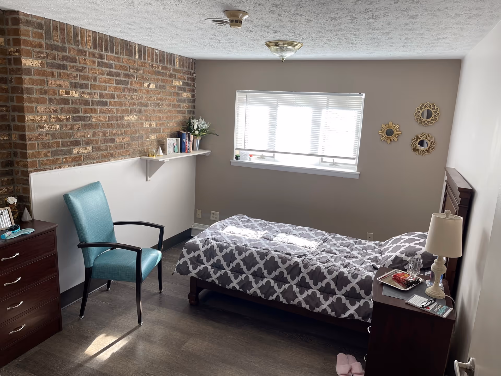 A small, cozy bedroom with a single bed covered in a gray and white patterned comforter. There is a wooden nightstand with a lamp, glass, and some items on it next to the bed. A teal armchair is positioned near a dark wooden dresser. The room has a brick accent wall with a white shelf holding books and a small plant. A window with blinds lets in natural light, and three decorative mirrors hang on the wall beside the window. The floor is dark wood, and a pair of pink slippers is on the floor near the nightstand.