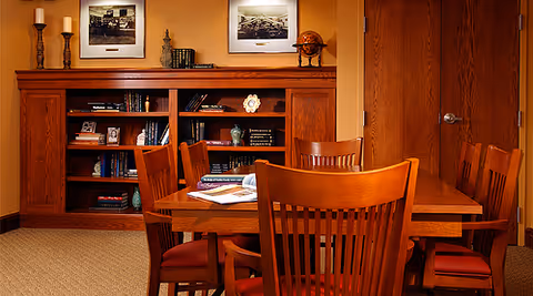 A cozy interior room featuring a wooden dining table surrounded by six wooden chairs with red cushions. Behind the table is a wooden bookshelf filled with books and decorative items, and above it are two framed black and white photographs. The walls are painted a warm orange color, and there is a closed wooden door to the right.