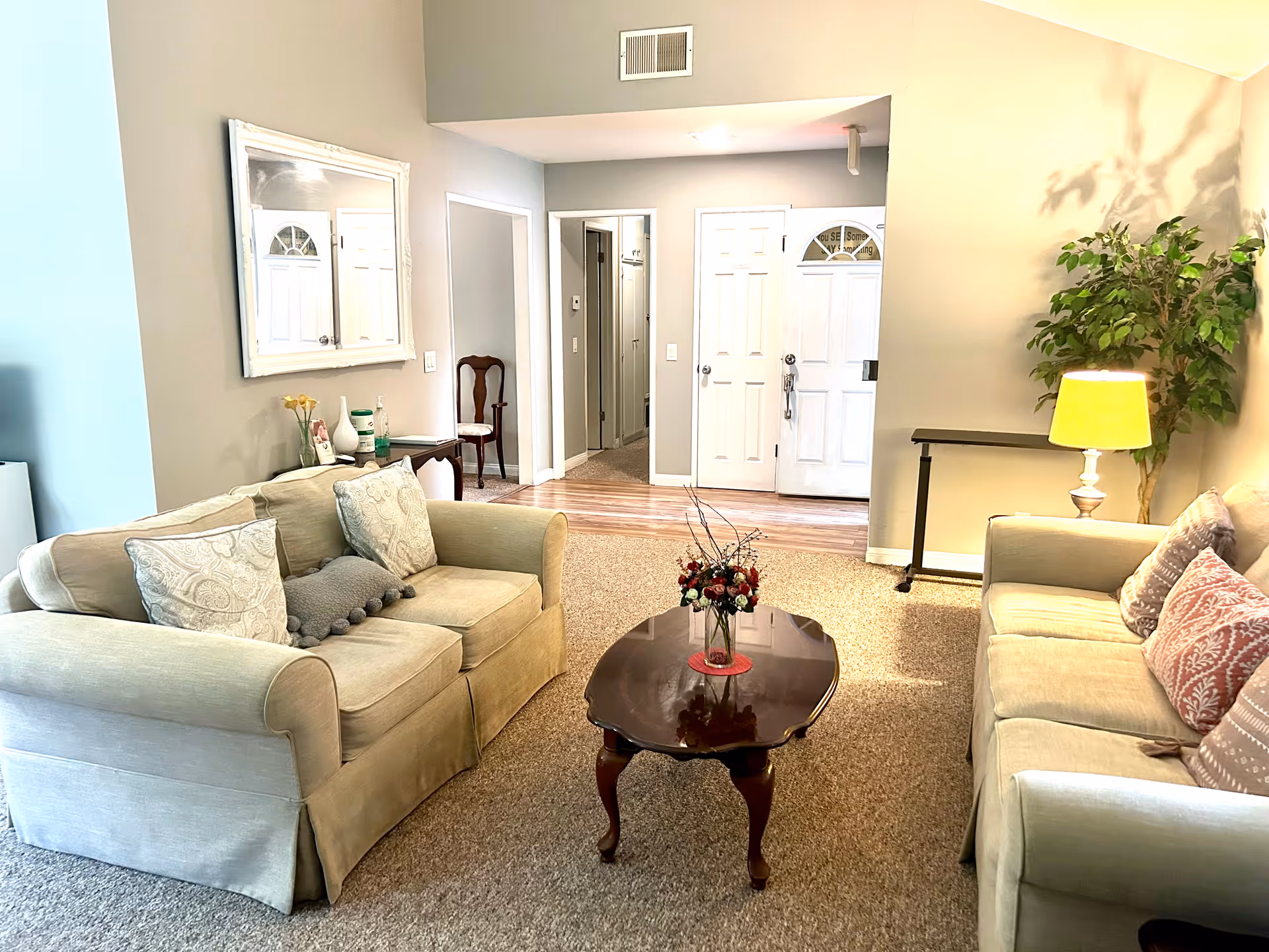 A cozy living room with two beige sofas facing each other, each adorned with decorative pillows. A dark wooden coffee table with a floral arrangement sits in the center on a carpeted floor. A large mirror hangs on the wall above a small console table with hand sanitizer and decorative items. In the background, there is a hallway with doors and a white front door with a small window. A floor lamp and a potted plant are positioned near one of the sofas.