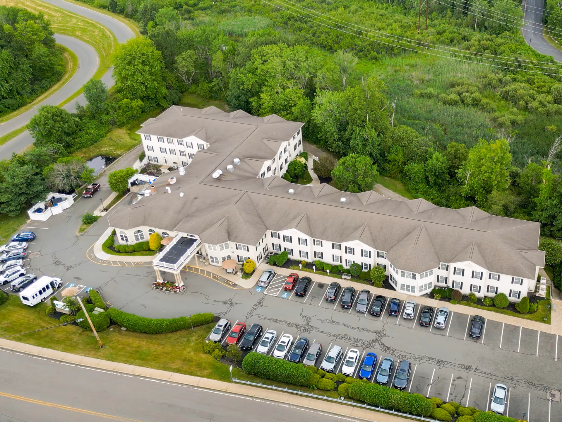 Aerial view of Benchmark Senior Living at Waltham Crossings, showing a large two-story building surrounded by greenery and trees. The building has a beige exterior with multiple windows and a covered entrance. There is a parking lot in front with several cars parked, and a curved road nearby.