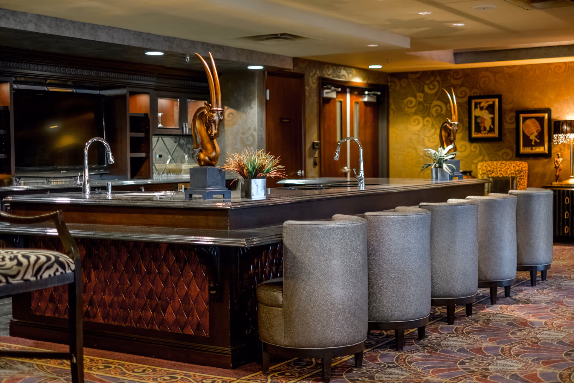 A luxurious interior space featuring a large dark wood bar counter with two sinks and tall chrome faucets. The bar is surrounded by six gray upholstered swivel chairs. Decorative elements include two bronze antelope head sculptures on the bar, a small plant, and framed artwork on the patterned golden wallpapered walls. The room has a richly patterned carpet and warm lighting.