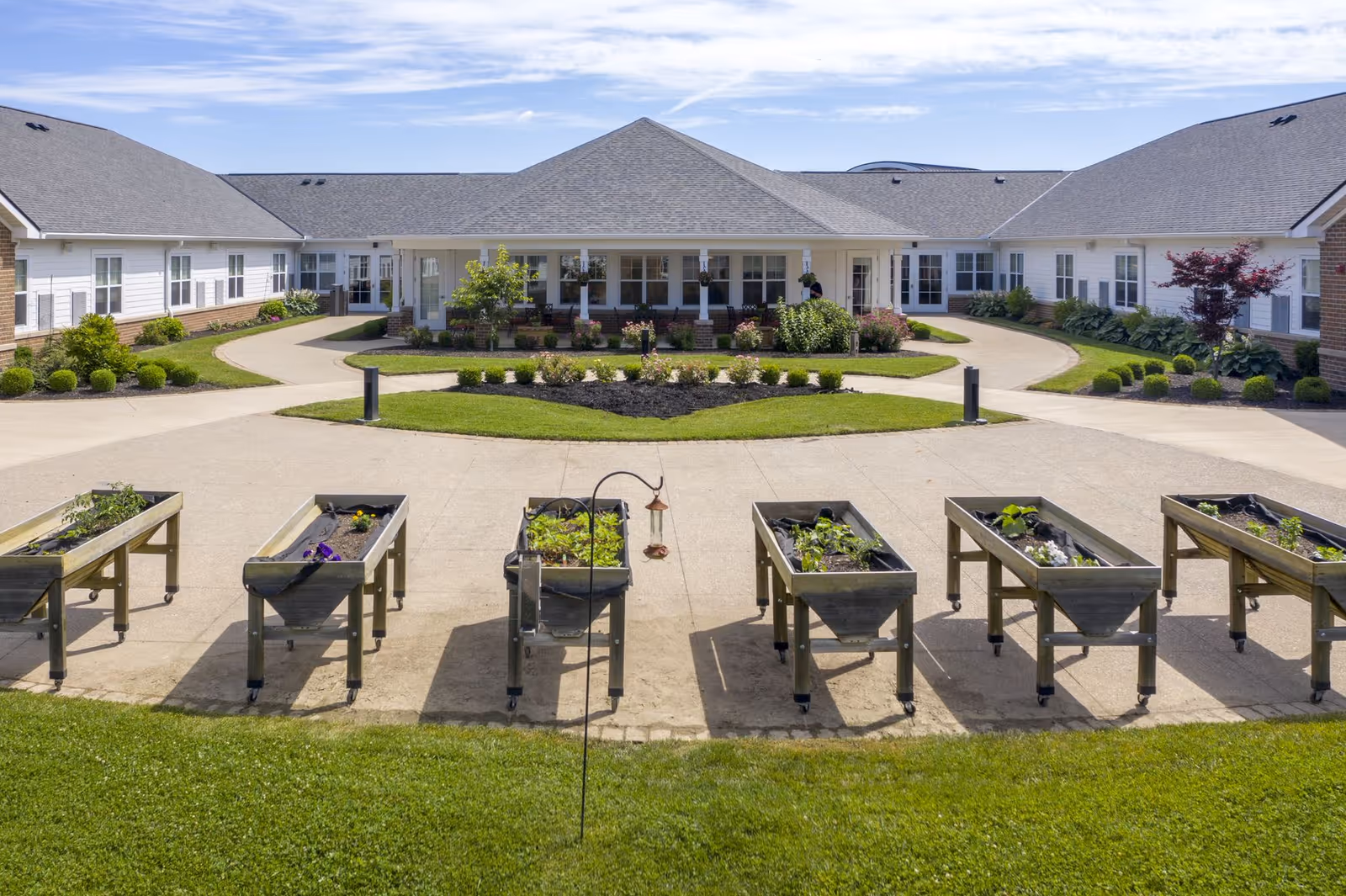 Outdoor view of a senior living facility with a paved courtyard featuring raised garden beds with plants. The building is single-story with a gray roof and white exterior walls, surrounded by well-maintained landscaping and green grass under a clear blue sky.