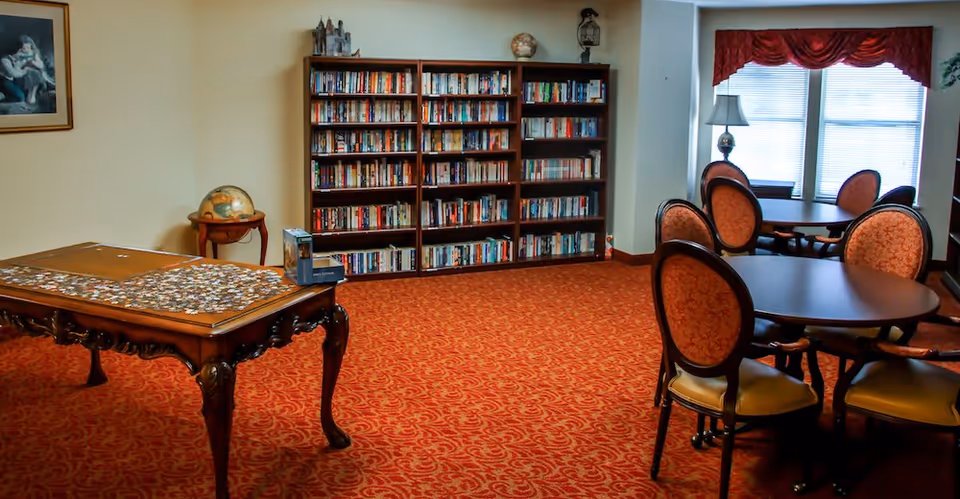 A cozy room with a large wooden table featuring a partially completed jigsaw puzzle, a globe on a small side table, a bookshelf filled with books, and round tables with upholstered chairs near a window with red valances.