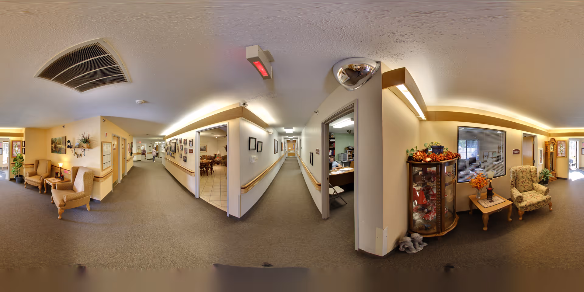 Panoramic interior view of a senior living facility corridor and common seating area with chairs, a display cabinet, and office window.