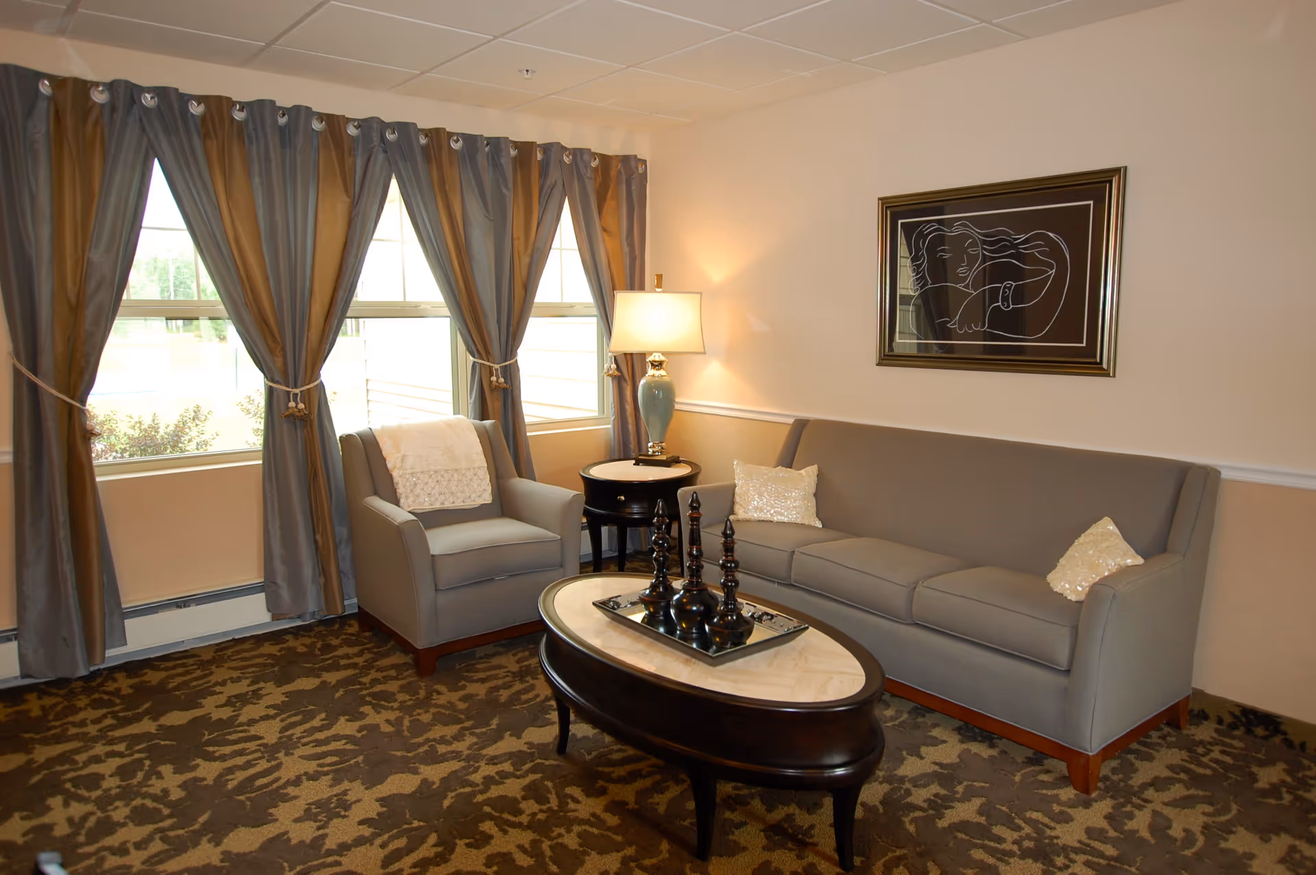 A cozy living room with a gray sofa and matching armchair, both adorned with light-colored decorative pillows. A dark wooden oval coffee table with a marble top holds three black decorative chess pieces on a tray. Next to the sofa is a round side table with a blue ceramic lamp. The room has large windows with gray curtains tied back, and a framed abstract line drawing hangs on the beige wall.