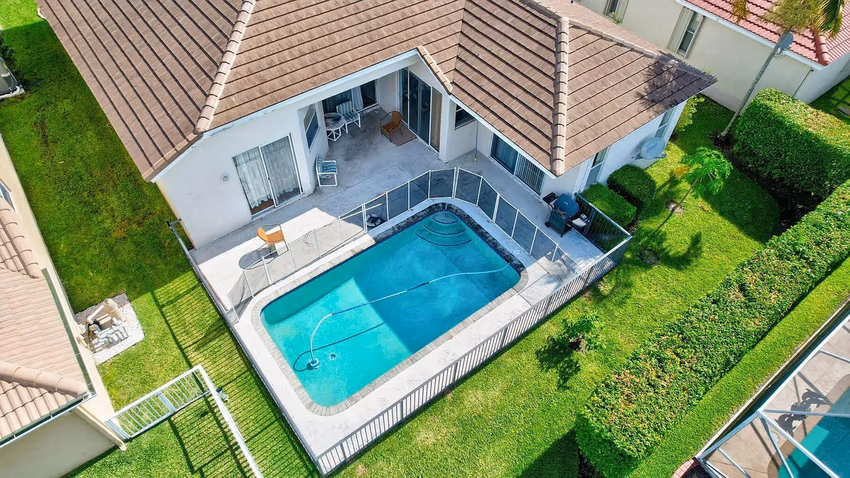 Aerial view of a single-story house with a fenced rectangular swimming pool and surrounding green lawn and patio.