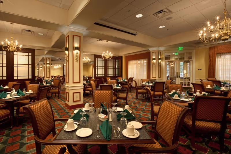 Elegant dining room with multiple set tables, upholstered chairs, chandeliers, and green napkins.