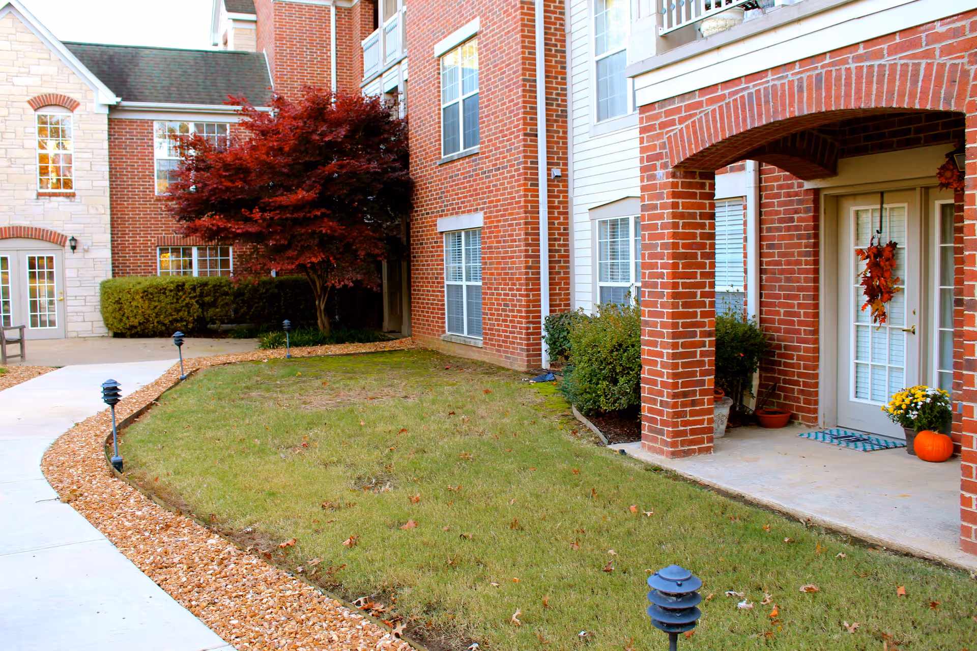 Outdoor view of a senior living facility with a brick building, a small grassy area, a curved concrete walkway lined with small black garden lights, and a tree with red leaves. The entrance to a unit is decorated with autumn-themed wreaths and a pumpkin.
