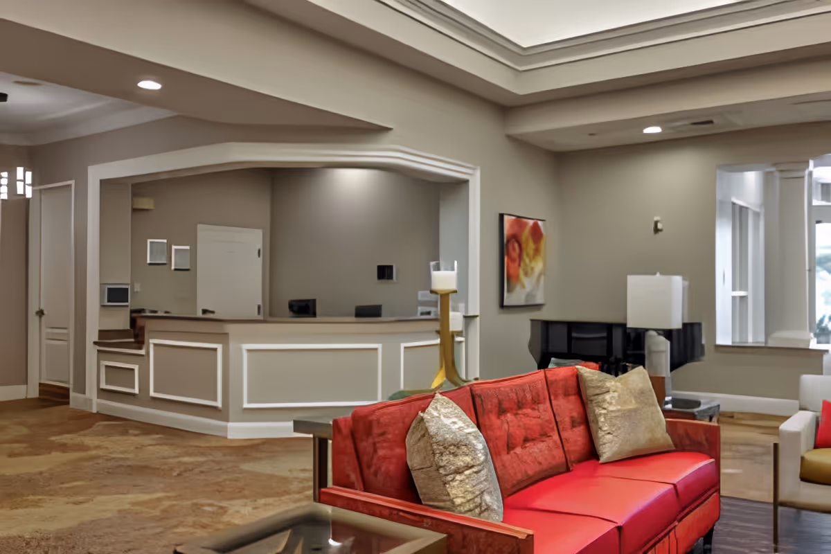 A spacious senior living facility common area with a red sofa adorned with gold pillows in the foreground. Behind the sofa is a reception desk with a computer and office chairs. The room features beige walls, a large window, a piano, a table lamp, and a colorful abstract painting on the wall.