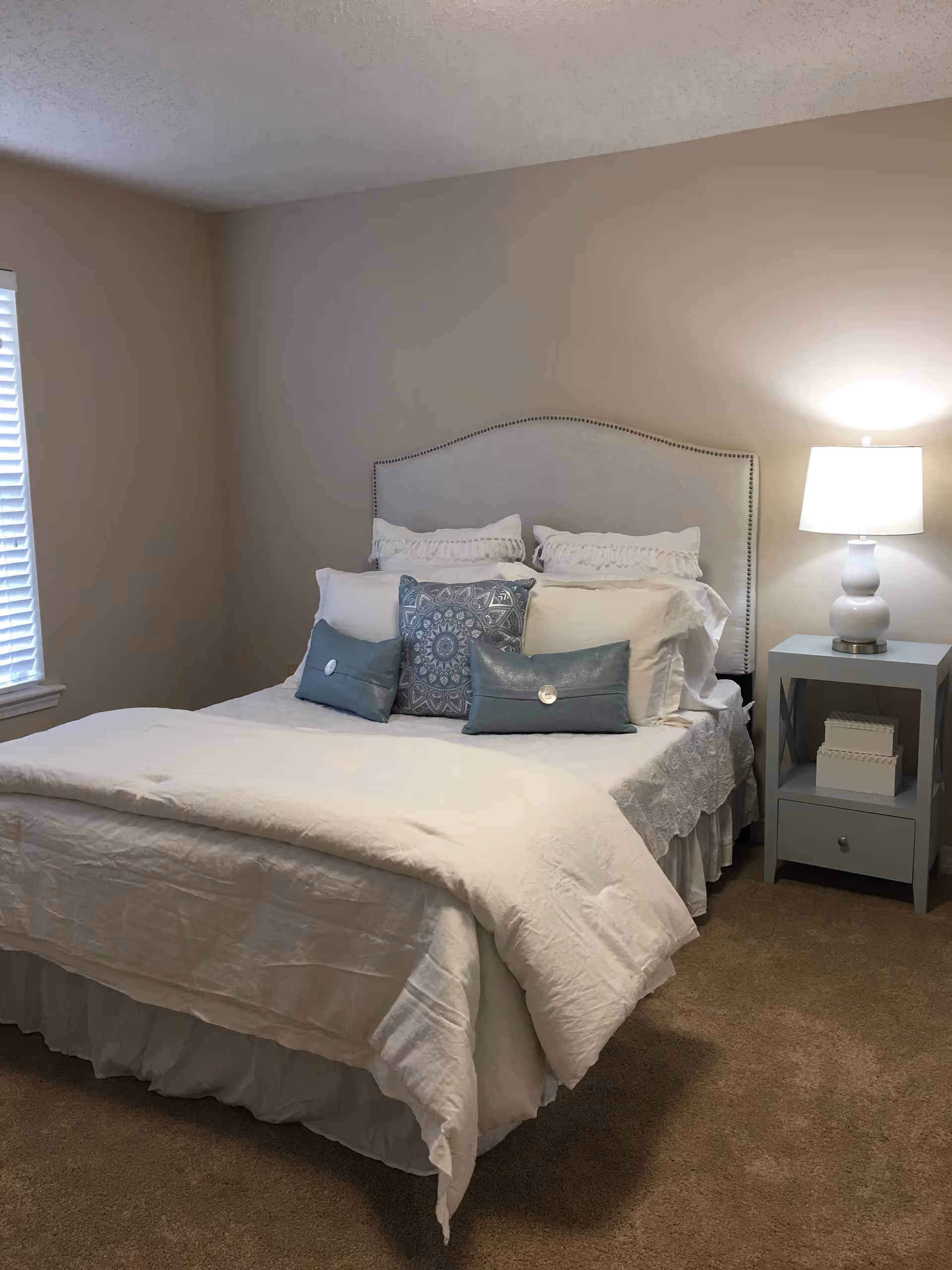 A neatly made bed with white bedding and decorative pillows in shades of blue and white, positioned against a beige wall. To the right of the bed is a light blue nightstand with a white lamp and two decorative boxes. A window with white blinds is visible on the left side of the room. The floor is carpeted in a light brown color.