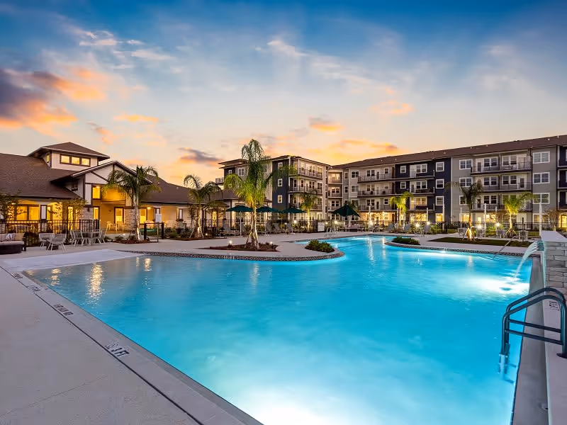 Outdoor swimming pool at a senior living facility during sunset, surrounded by palm trees, lounge chairs, umbrellas, and multi-story residential buildings in the background.