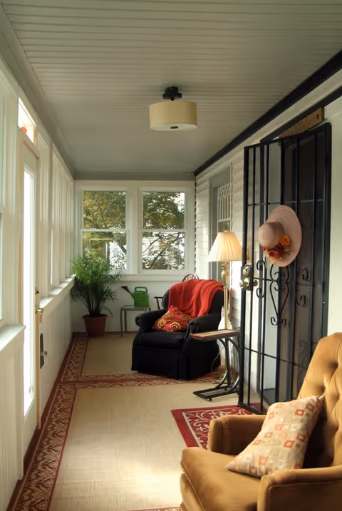 A cozy enclosed porch with large windows letting in natural light. The space features a black armchair with a red throw blanket and patterned pillow, a tan armchair with a decorative pillow, a floor lamp, a potted plant, and a watering can. A pink hat with flowers hangs on a black metal door gate.