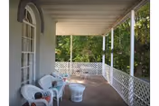 Covered porch area with white lattice railing and white wicker chairs with cushions arranged around a small white wicker table. The porch overlooks a green, leafy outdoor area.