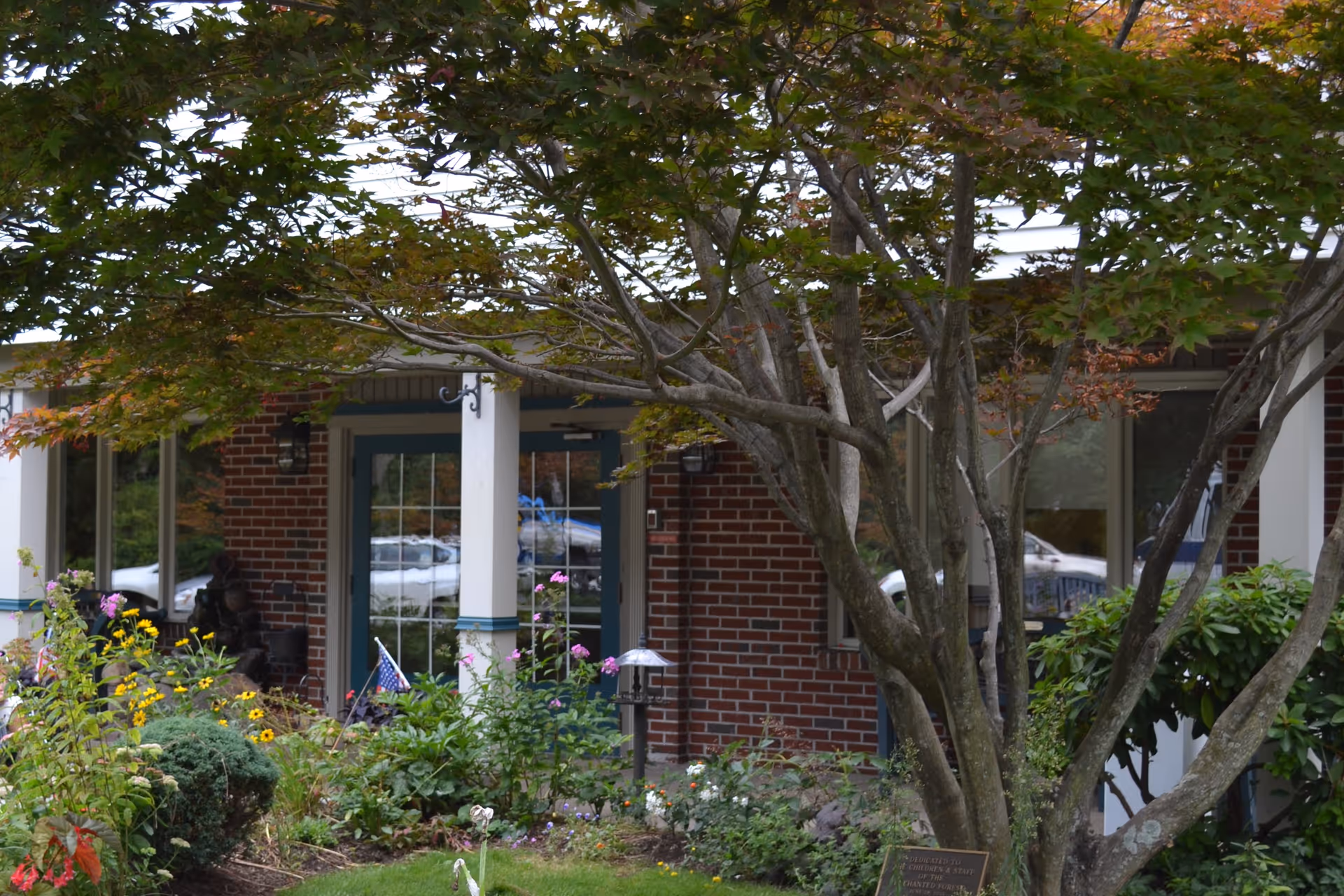 Front entrance of a brick building partially obscured by a large tree with green and orange leaves. There are various plants and flowers in a garden bed in front of the building, and a glass door with white framing is visible. A small American flag is planted among the flowers.