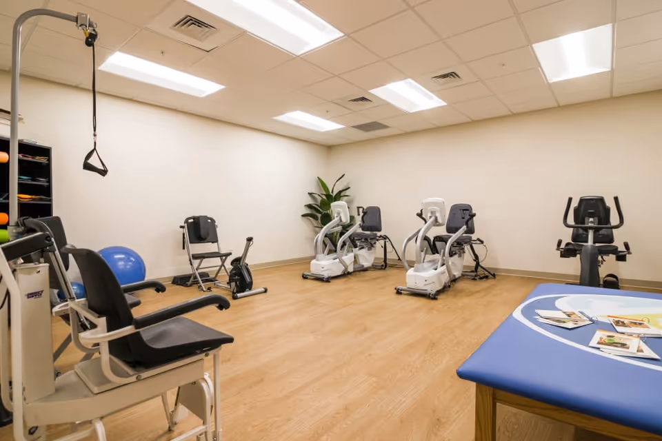 Bright exercise/therapy room with several recumbent exercise bikes, a stationary bike, therapy chairs, an exercise ball, and a padded treatment table.