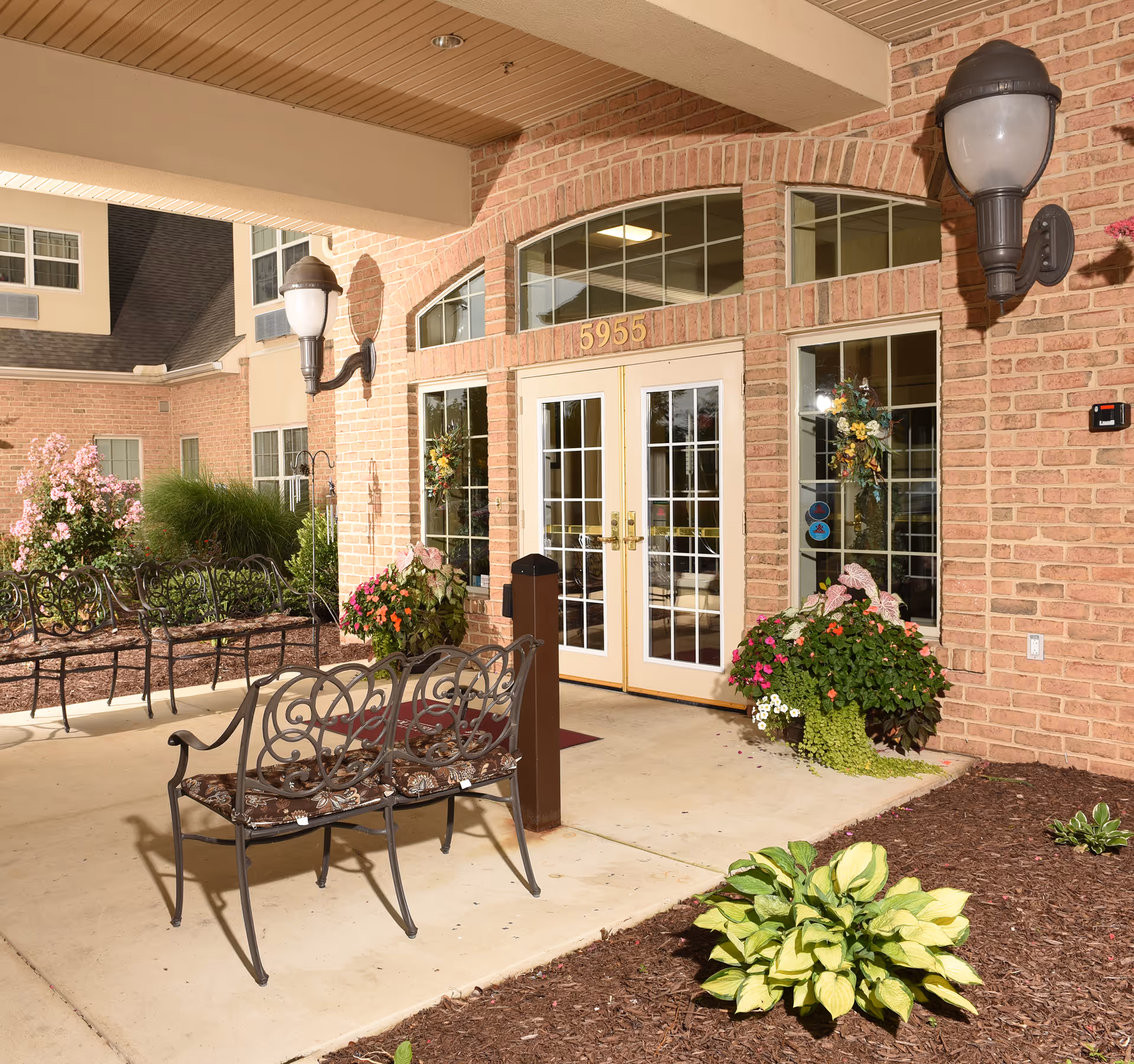 Entrance of a brick building with double glass doors numbered 5955, flanked by large windows decorated with floral wreaths. There are two black metal benches with cushions on a concrete patio, surrounded by landscaped flower beds with various plants and flowers. Two wall-mounted outdoor lamps are visible on either side of the entrance.