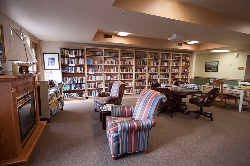 A cozy community room with a large bookshelf filled with books along the back wall. Two striped armchairs are positioned near a wooden fireplace with a decorative sailboat on the mantel. There is a round table with chairs and another table with chairs in the background. The room has carpeted flooring and soft lighting.