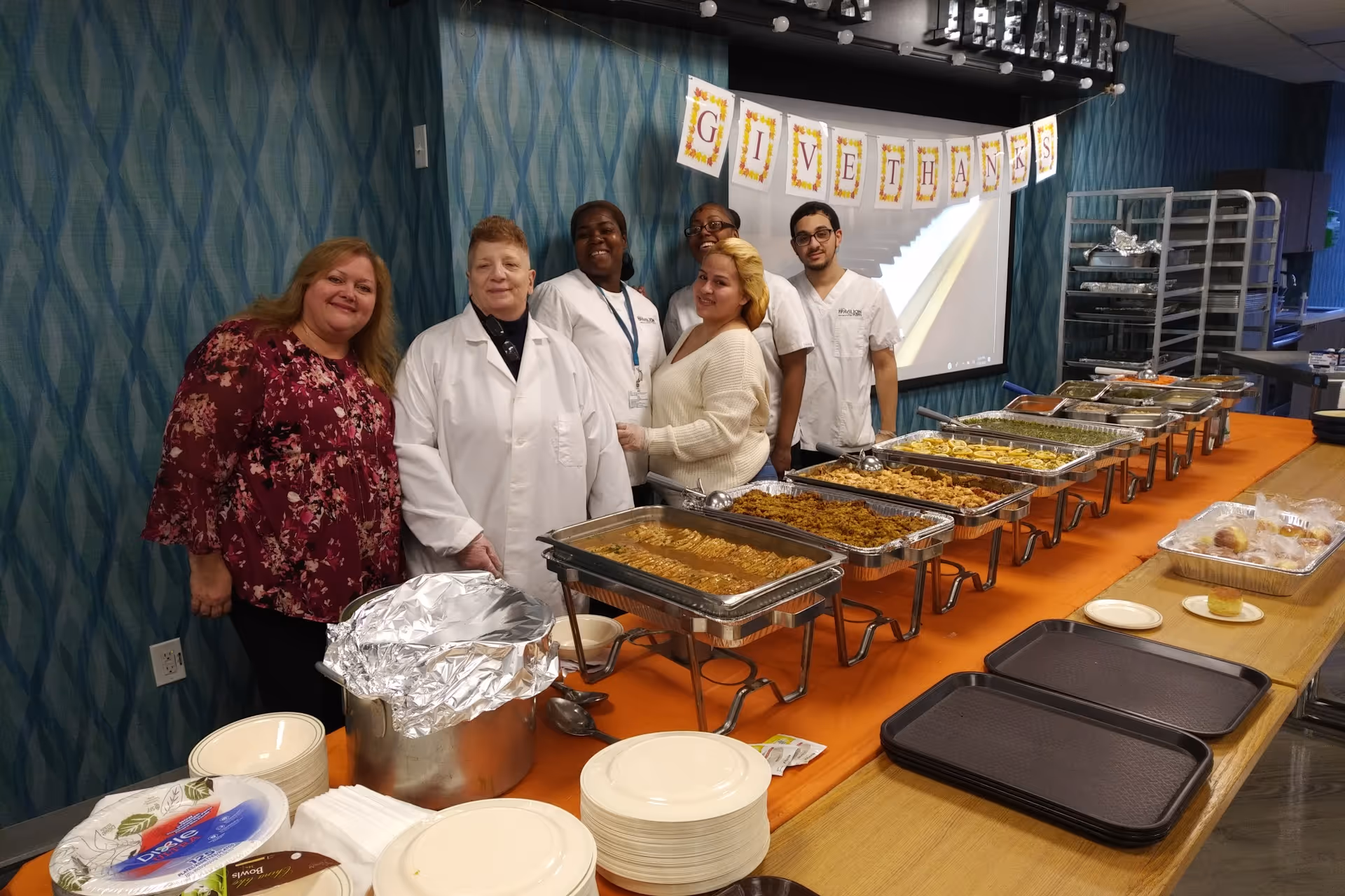Five staff members standing behind a long table filled with trays of food in a dining area decorated with a 'Give Thanks' banner.