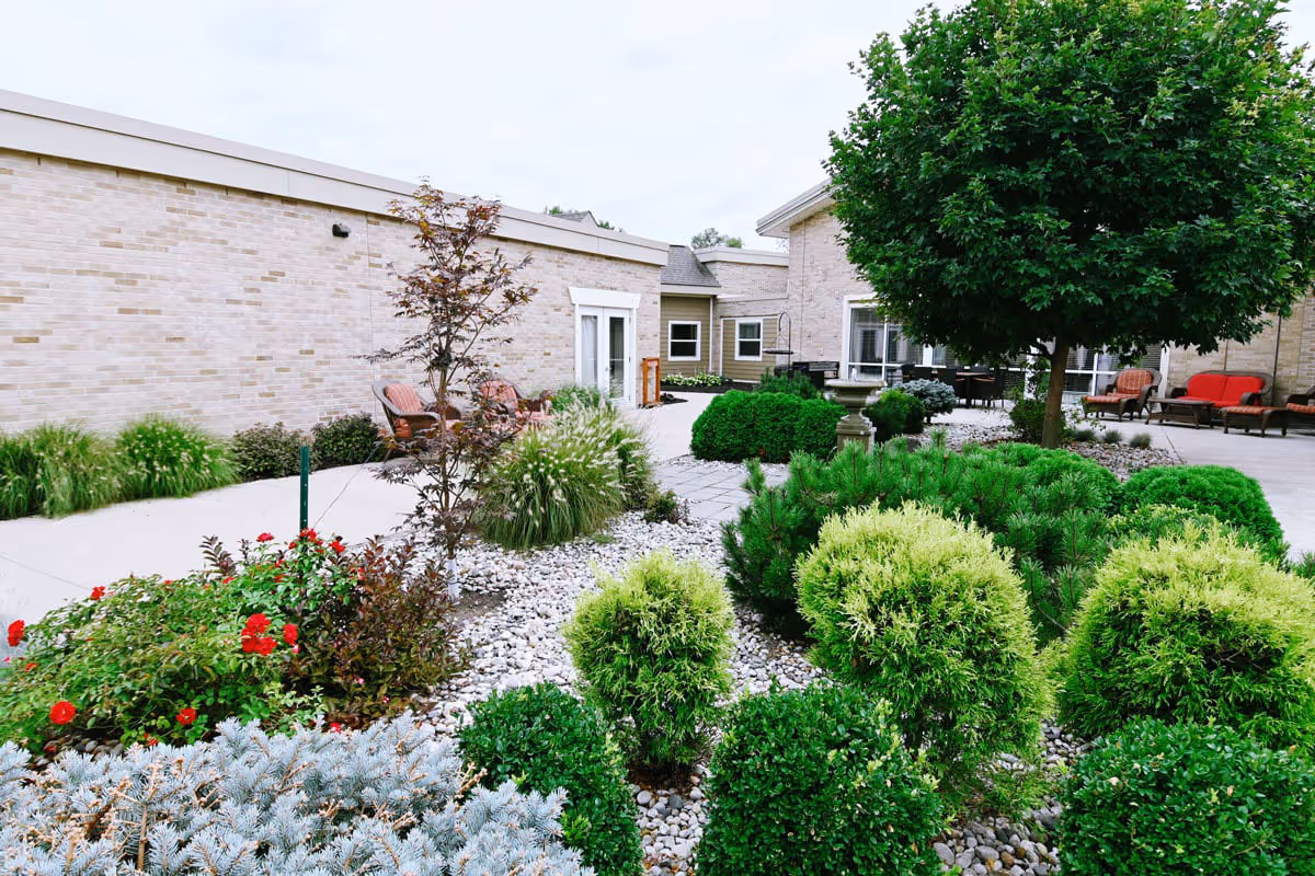 A landscaped outdoor courtyard area at The Cortland Wyoming featuring various green shrubs, small trees, and flowering plants surrounded by a stone and concrete pathway. There are several seating areas with chairs and a red cushioned bench near the building walls.