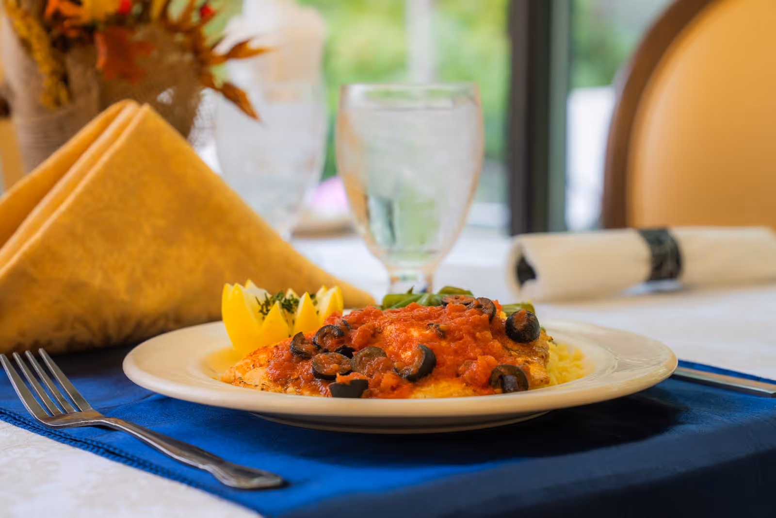 A close-up of a plated meal featuring a dish topped with tomato sauce and sliced black olives, garnished with lemon wedges and herbs, set on a white plate with a blue placemat. In the background, there is a glass of water, a folded yellow napkin, and a rolled white napkin with a black napkin ring on a dining table near a window.