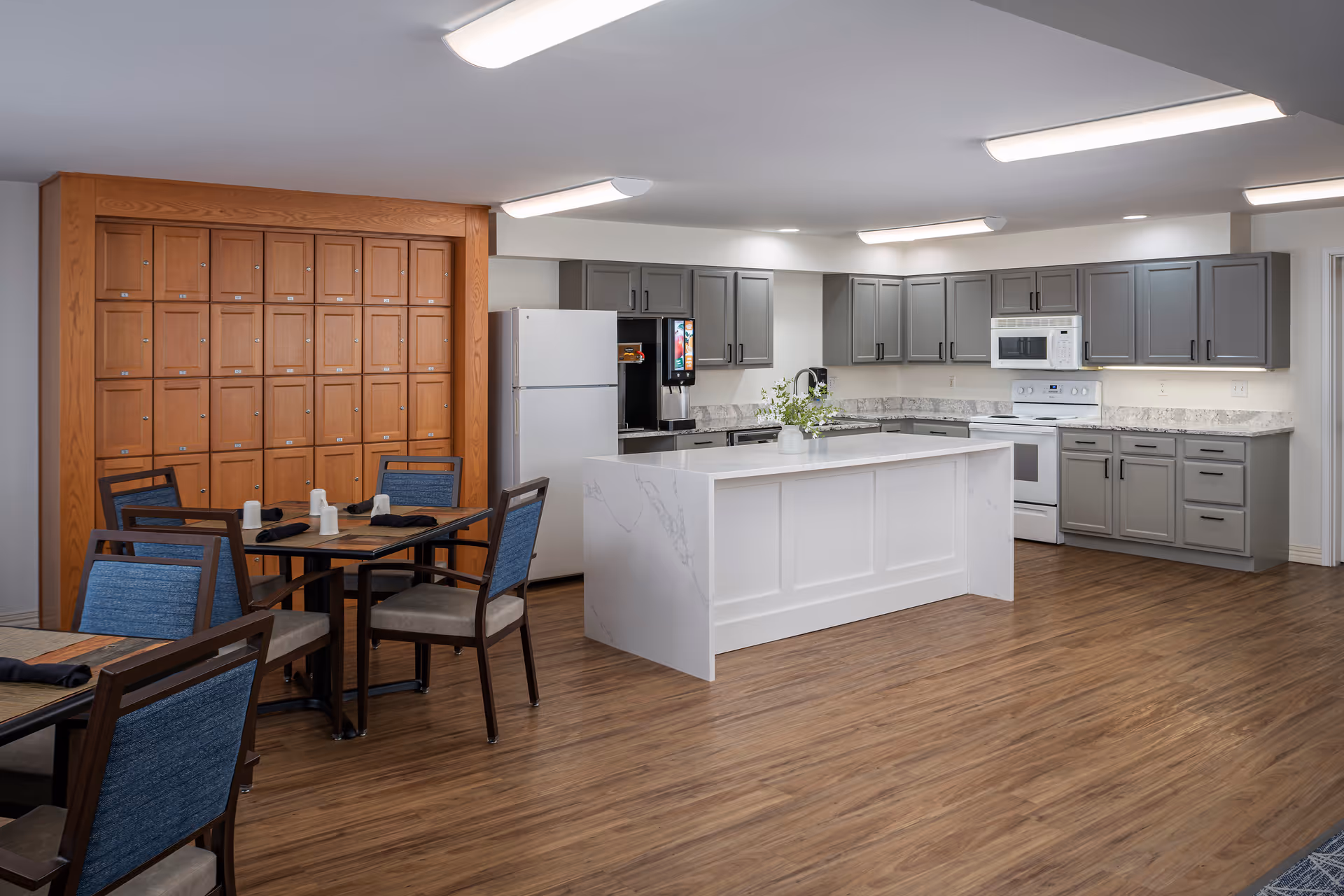 A modern senior living facility kitchen and dining area with gray cabinets, white countertops, a white kitchen island, a refrigerator, microwave, and a coffee machine. There are wooden mailboxes on the wall and dining tables with blue cushioned chairs set with black napkins and white cups. The floor is wood, and the ceiling has bright rectangular lights.