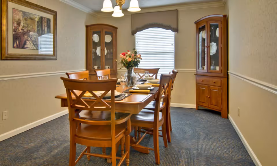 A dining room with a wooden table set for six people, featuring plates, glasses, and napkins. There are two wooden china cabinets in the corners, a window with blinds and a valance, a framed painting on the wall, and a ceiling light fixture above the table.