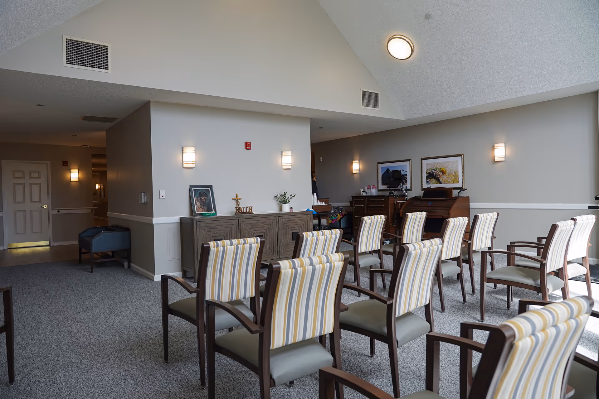 Interior room with multiple chairs arranged in rows facing a wall with framed pictures and a wooden cabinet. The room has beige walls, carpeted floor, and ceiling lights. There is a hallway visible to the left with a closed door and additional seating.
