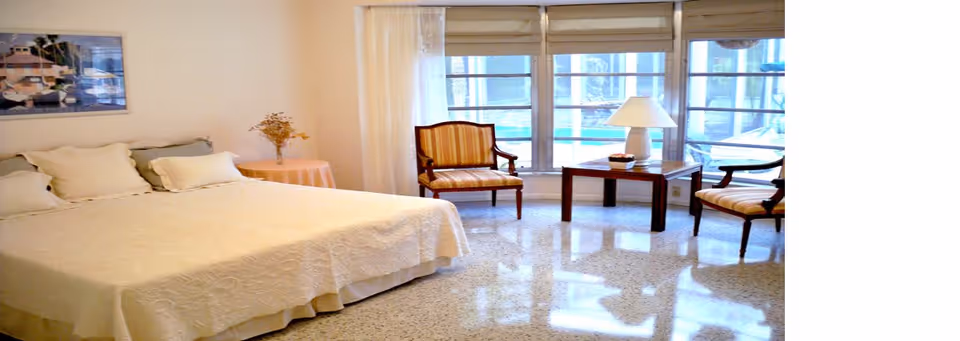 Bright bedroom with a large bed, two armchairs, a side table and large windows overlooking an outdoor area.